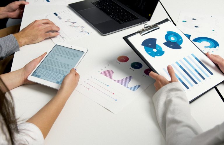 Zwei Personen analysieren Diagramme und Daten auf einem Tisch mit Laptop und Tablet.